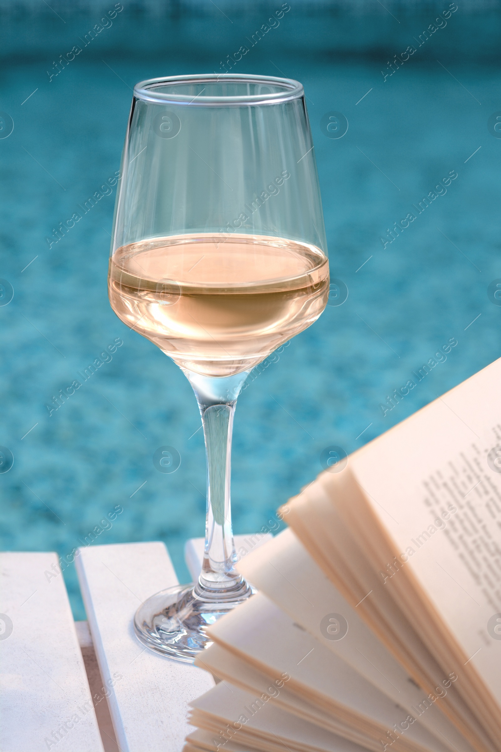 Photo of Glass of tasty wine and open book on wooden table near swimming pool