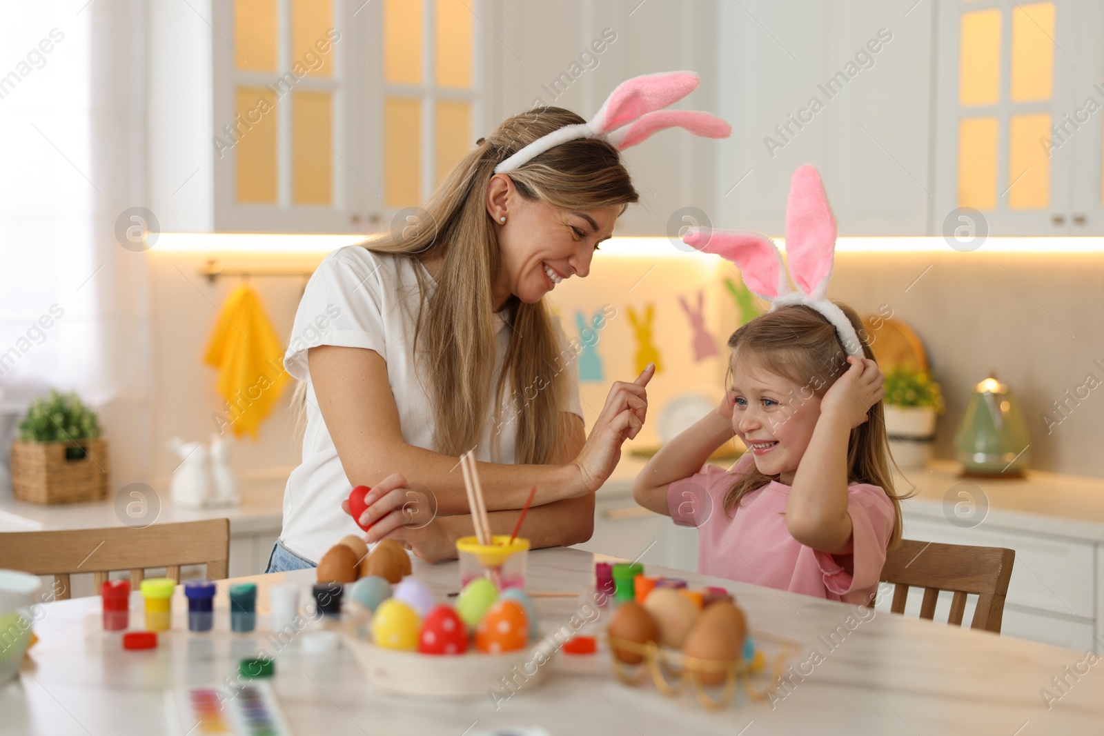 Photo of Easter celebration. Mother with her cute daughter having fun while painting eggs at white marble table in kitchen