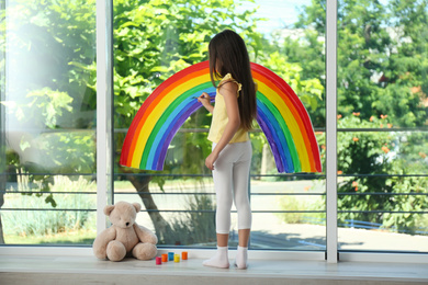 Photo of Little girl drawing rainbow on window with paints indoors. Stay at home concept