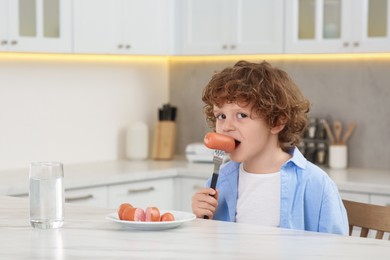 Photo of Cute little boy eating sausages at table in kitchen