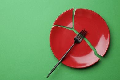 Photo of Pieces of broken ceramic plate and fork on green background, flat lay. Space for text