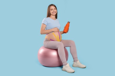 Pregnant woman with kinesio tapes holding water bottle on fitball against light blue background