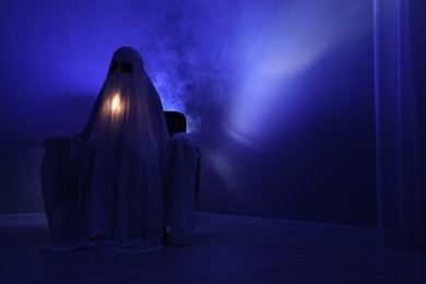Creepy ghost. Woman covered with sheet sitting in armchair in blue light, space for text