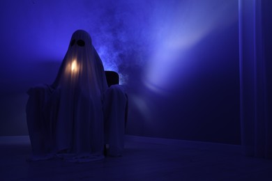 Photo of Creepy ghost. Woman covered with sheet sitting in armchair in blue light, space for text