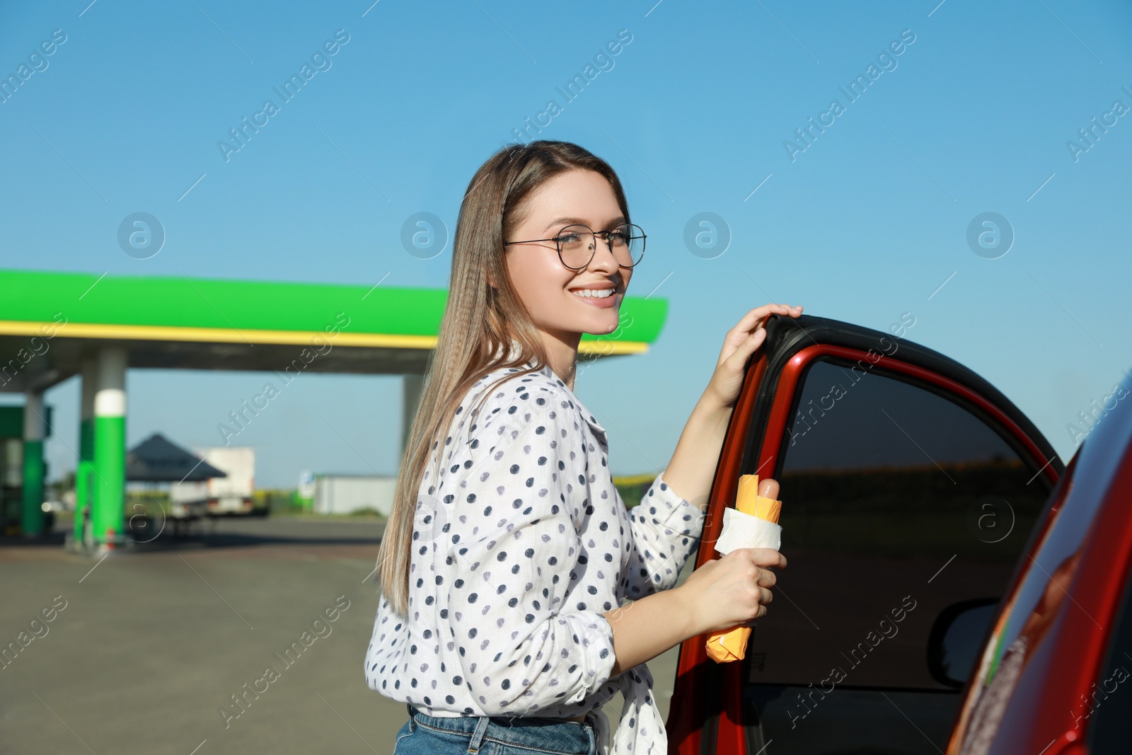 Photo of Beautiful young woman with hot dog opening car door at gas station