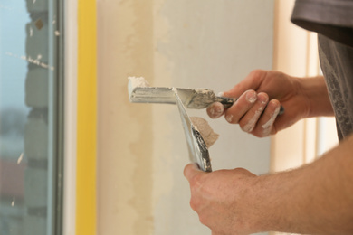 Photo of Professional worker with putty knifes and plaster near window indoors, closeup. Interior repair