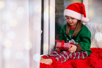 Cute little girl in Santa hat holding Christmas present on window sill at home