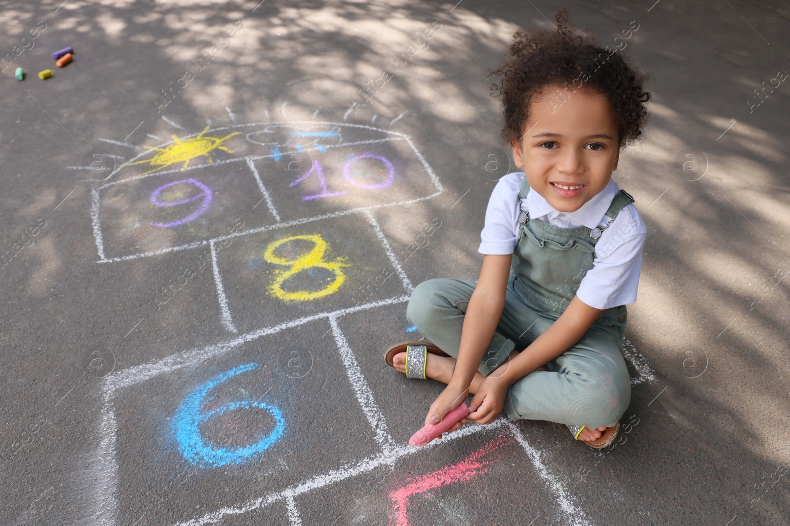 Photo of Little African American girl drawing hopscotch with chalk on asphalt outdoors. Happy childhood