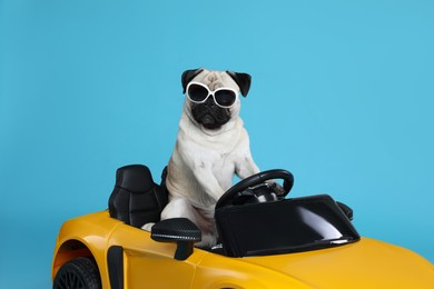 Funny pug dog with sunglasses in toy car on light blue background