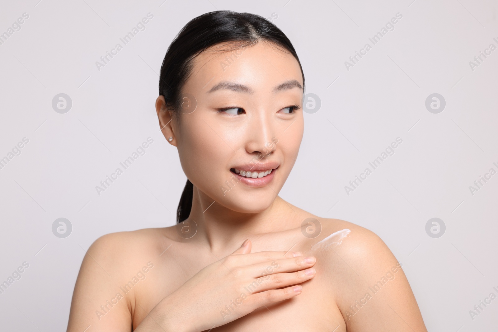 Photo of Beautiful young Asian woman applying body cream onto shoulder on light grey background