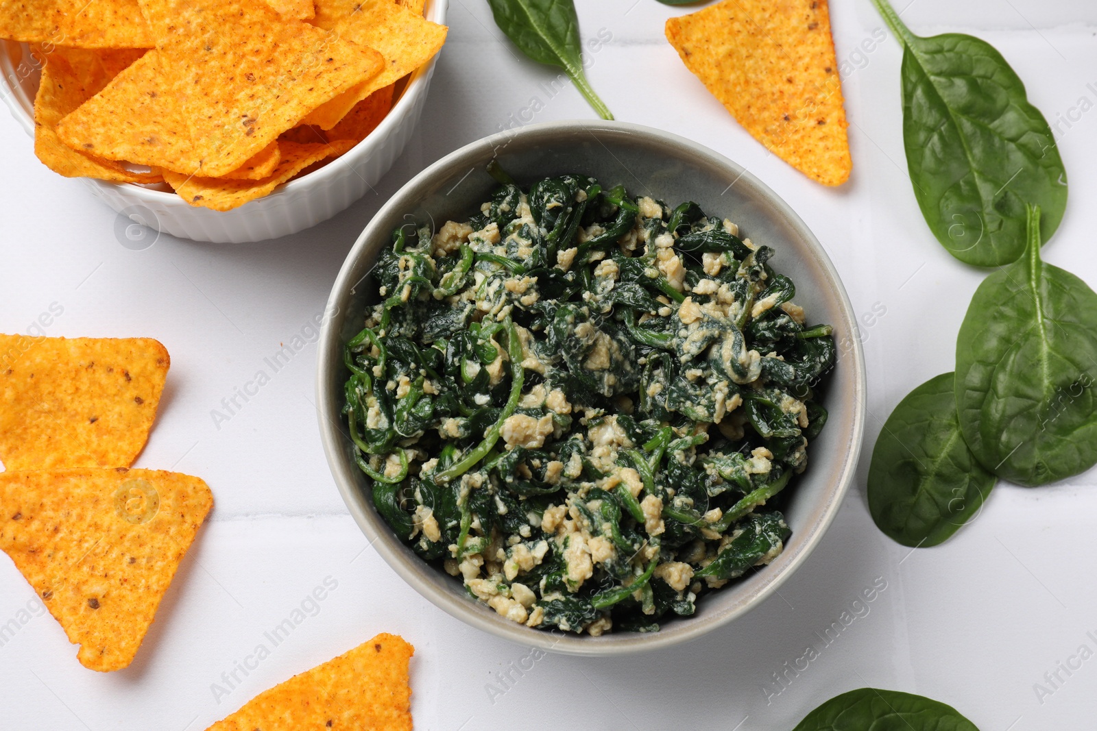 Photo of Tasty spinach dip with egg in bowl and nachos chips on white table, flat lay