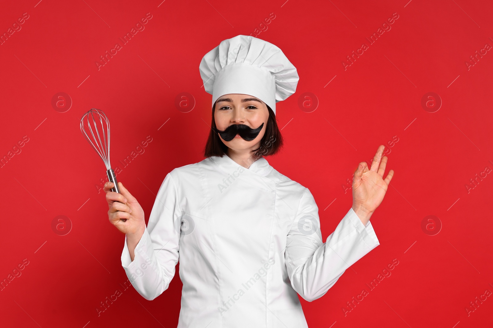 Photo of Confectioner with funny artificial moustache and whisk showing ok gesture on red background