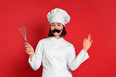 Photo of Confectioner with funny artificial moustache and whisk showing ok gesture on red background