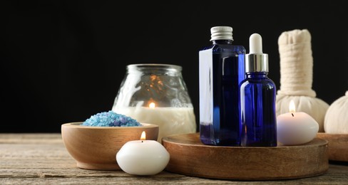 Spa composition. Cosmetic products, herbal bags, burning candles and sea salt on wooden table against black background, space for text