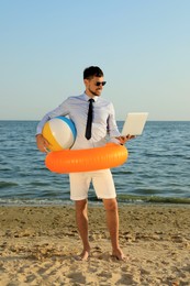 Happy man with inflatable ring, ball and laptop near sea on beach. Business trip