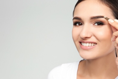 Young woman plucking eyebrow with tweezers on light background