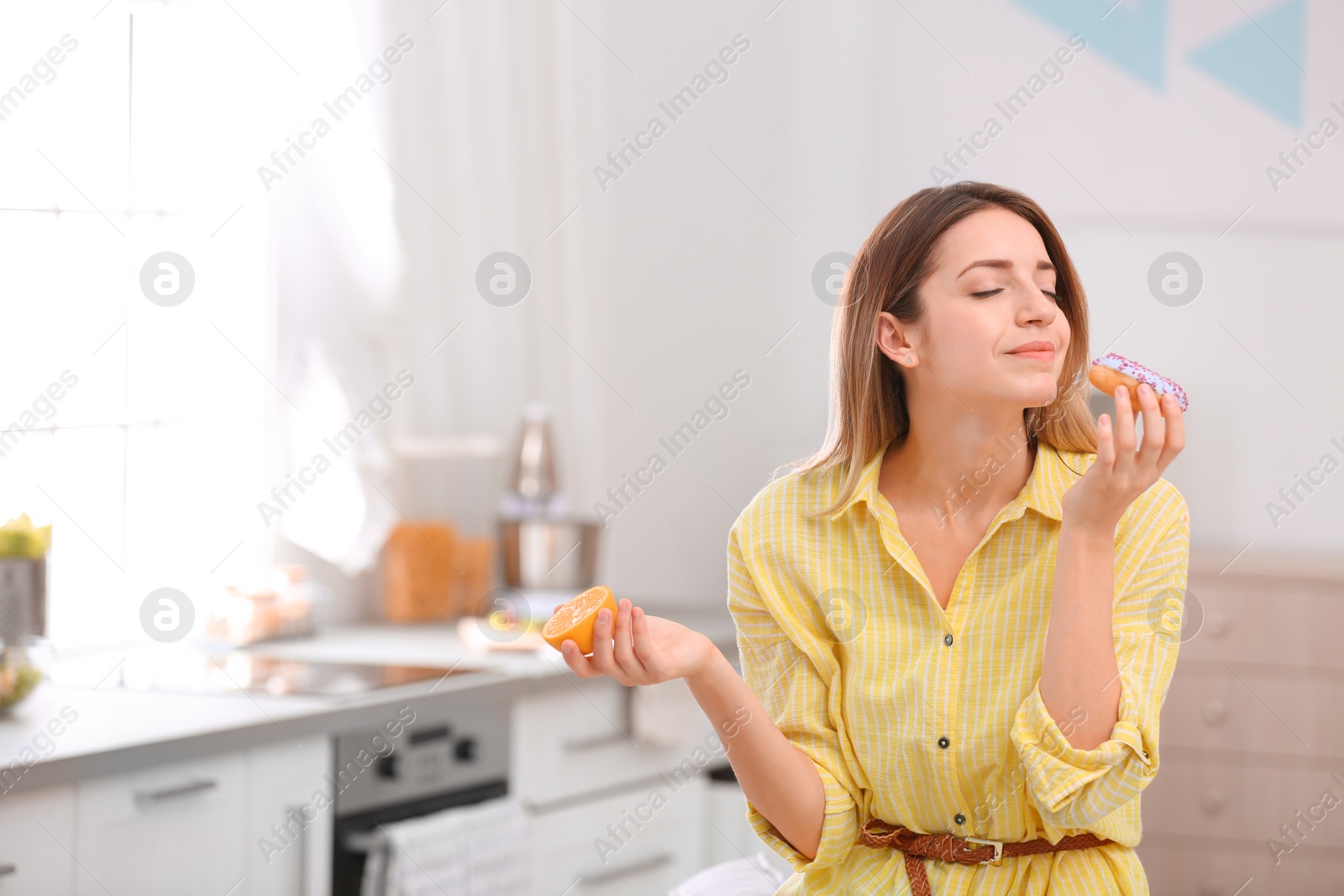 Photo of Young woman choosing between orange and donut in kitchen, space for text. Healthy diet