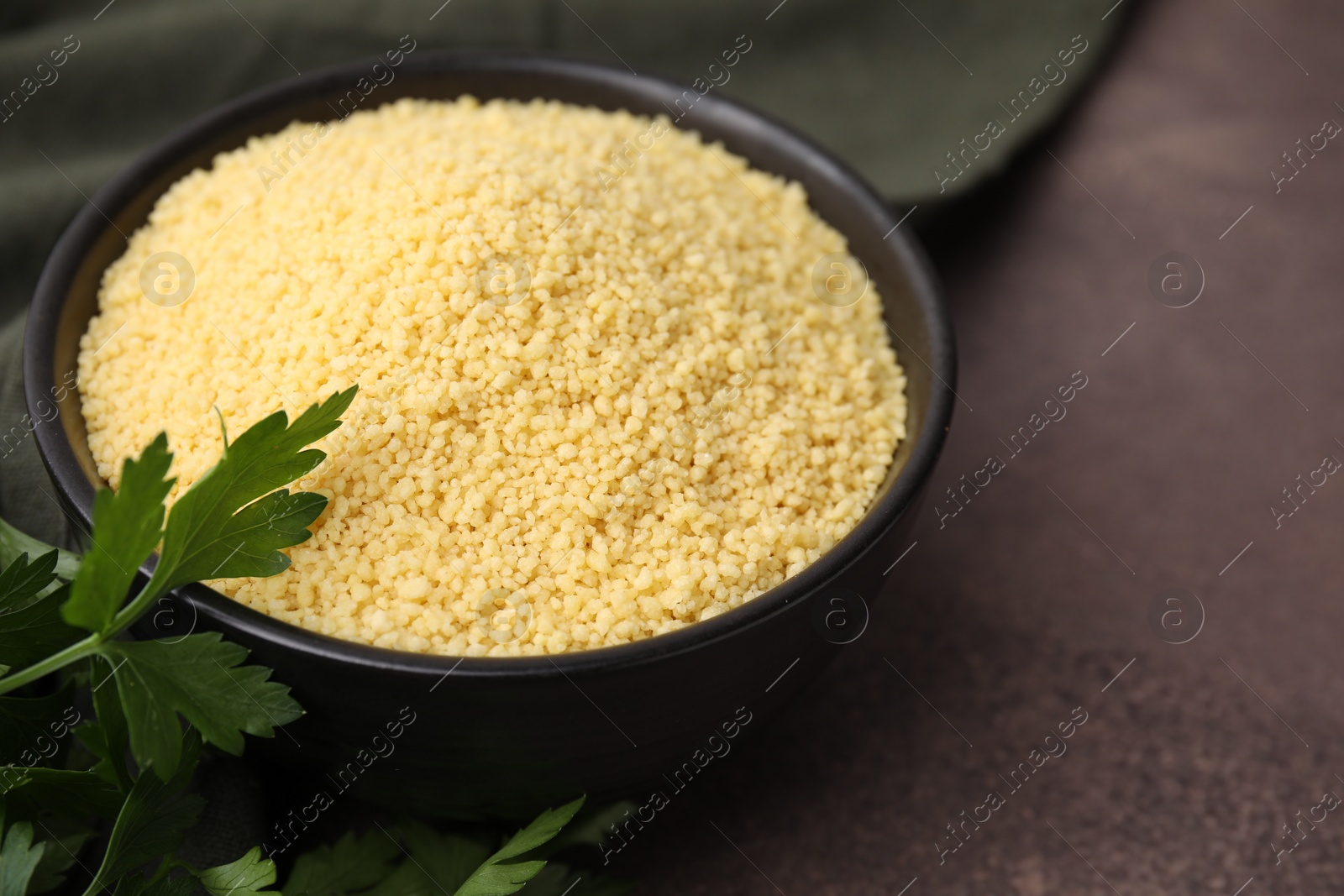 Photo of Raw couscous in bowl and parsley on brown table, closeup