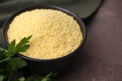 Photo of Raw couscous in bowl and parsley on brown table, closeup