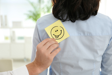 Photo of Man putting funny smiling face sticker onto colleague's back in office, closeup. April fool's day