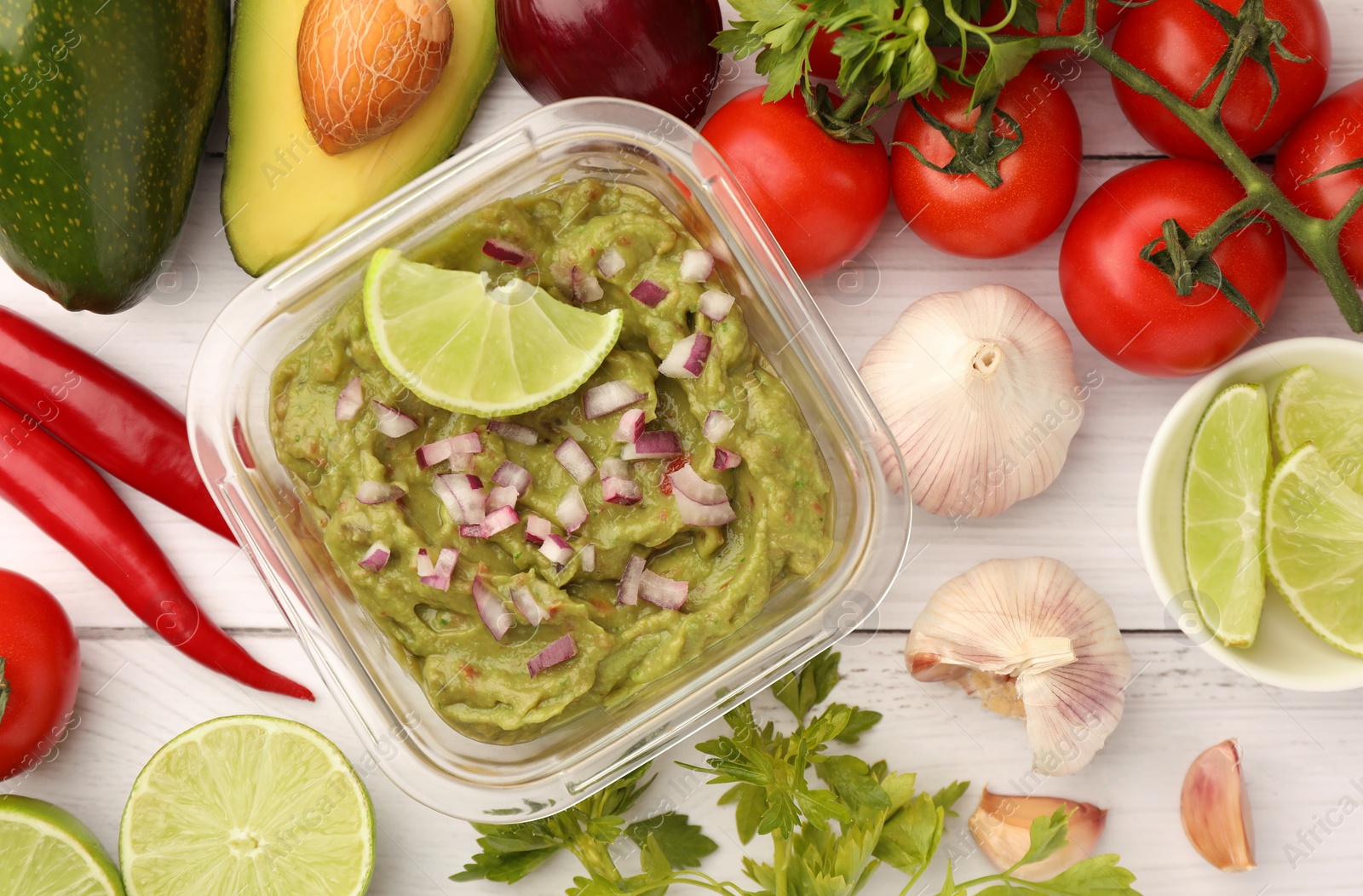 Photo of Bowl of delicious guacamole with onion, lime and ingredients on white wooden table, flat lay