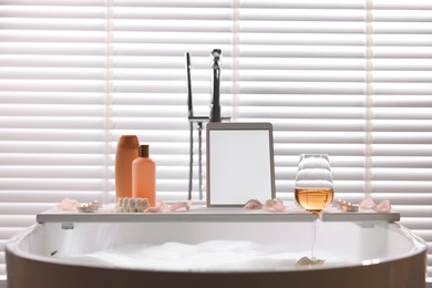 Photo of Wooden tray with tablet, wine, toiletries and flower petals on bathtub in bathroom