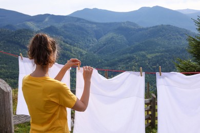 Photo of Woman hanging clean laundry with clothespins on washing line in mountains, back view