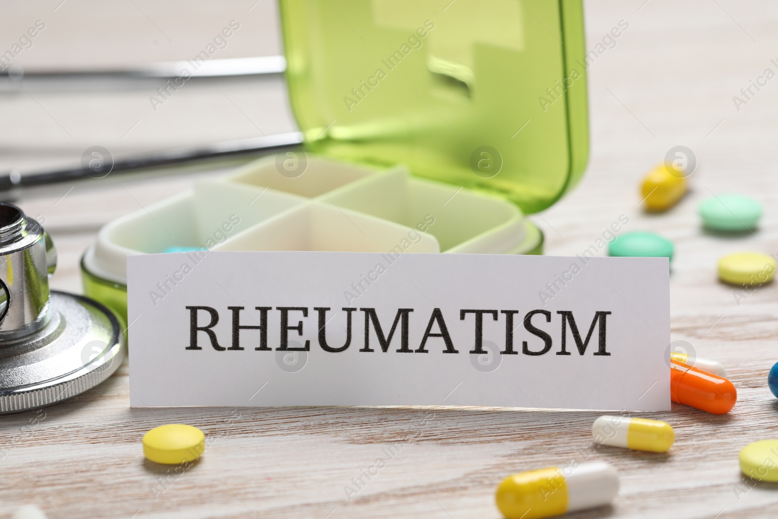 Photo of Card with word Rheumatism, stethoscope and pills on white wooden table, closeup