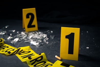 Broken bottle, yellow tape and evidence markers on black slate table, closeup. Crime scene