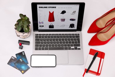 Photo of Online store website on laptop screen. Computer, smartphone, credit cards, women's shoes, stationery and lipstick on white background, above view