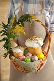 Photo of Woman holding basket with traditional Easter cakes, dyed eggs and flowers indoors, closeup