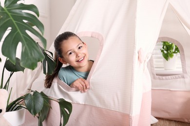 Photo of Cute little girl peeking out window of toy wigwam at home
