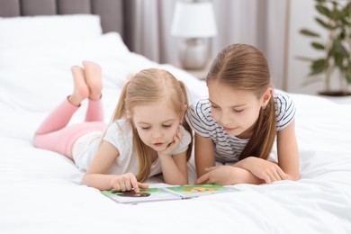 Photo of Cute little sisters reading book together on bed at home