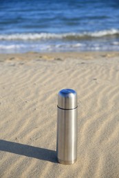 Metallic thermos with hot drink on sand near sea