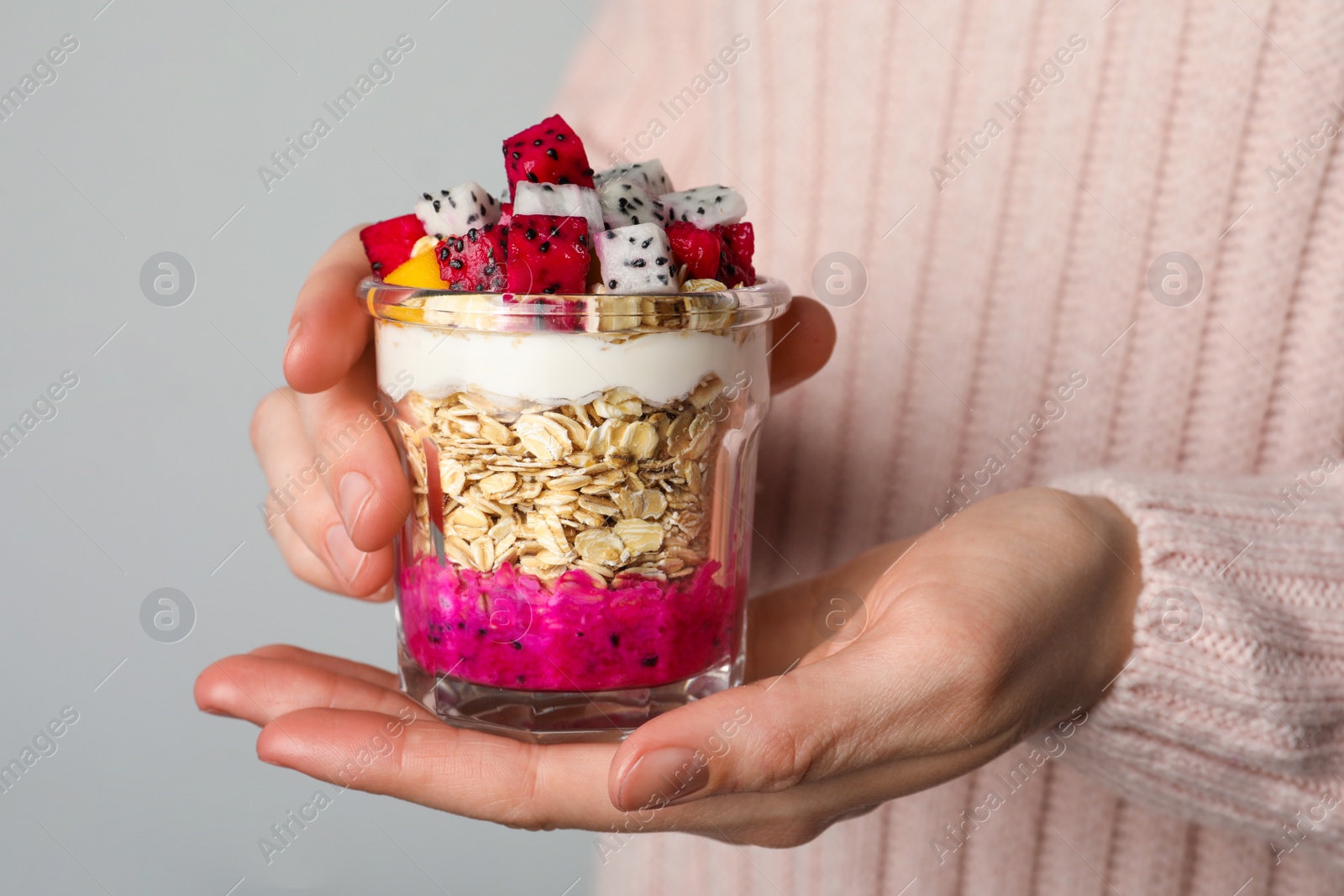 Photo of Woman holding glass of granola with pitahaya, yogurt and mango on grey background, closeup