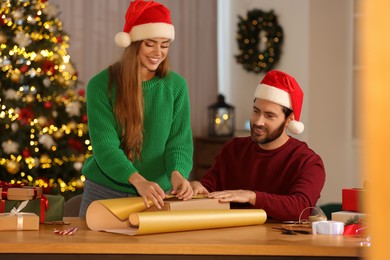 Happy couple in Santa hats decorating Christmas gift with wrapping paper at table in room