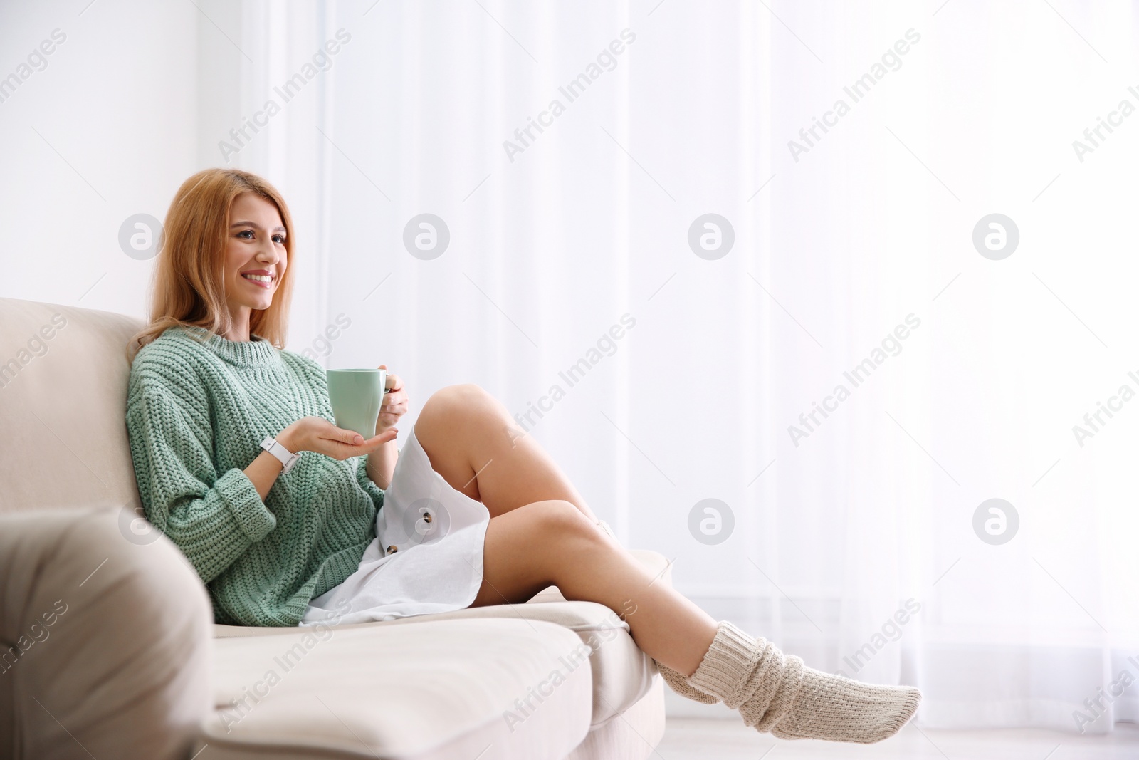 Photo of Young woman with cup of drink relaxing on couch near window at home. Space for text