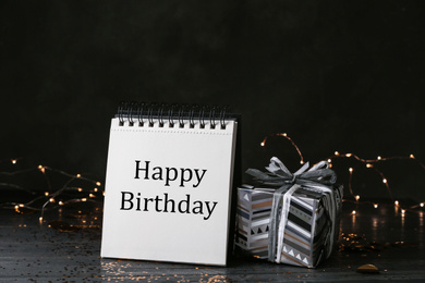 Photo of Notepad with greeting HAPPY BIRTHDAY and gift on wooden table
