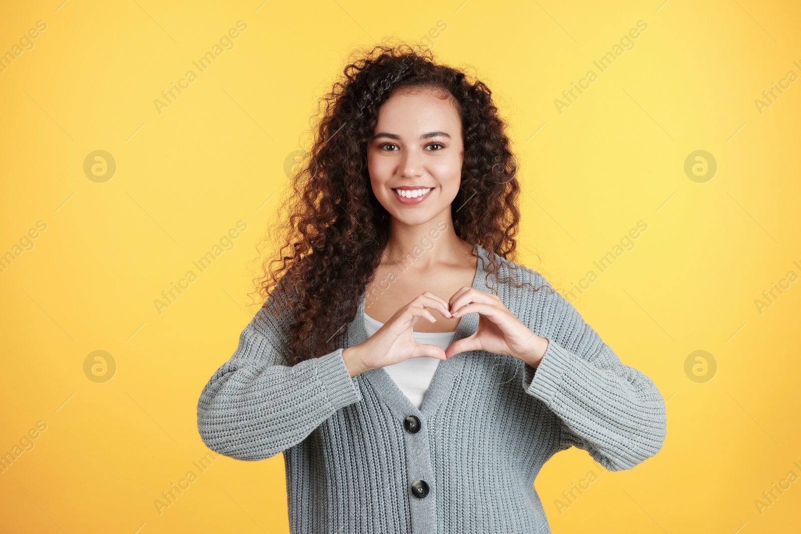 Photo of Happy young African-American woman making heart with hands on yellow background