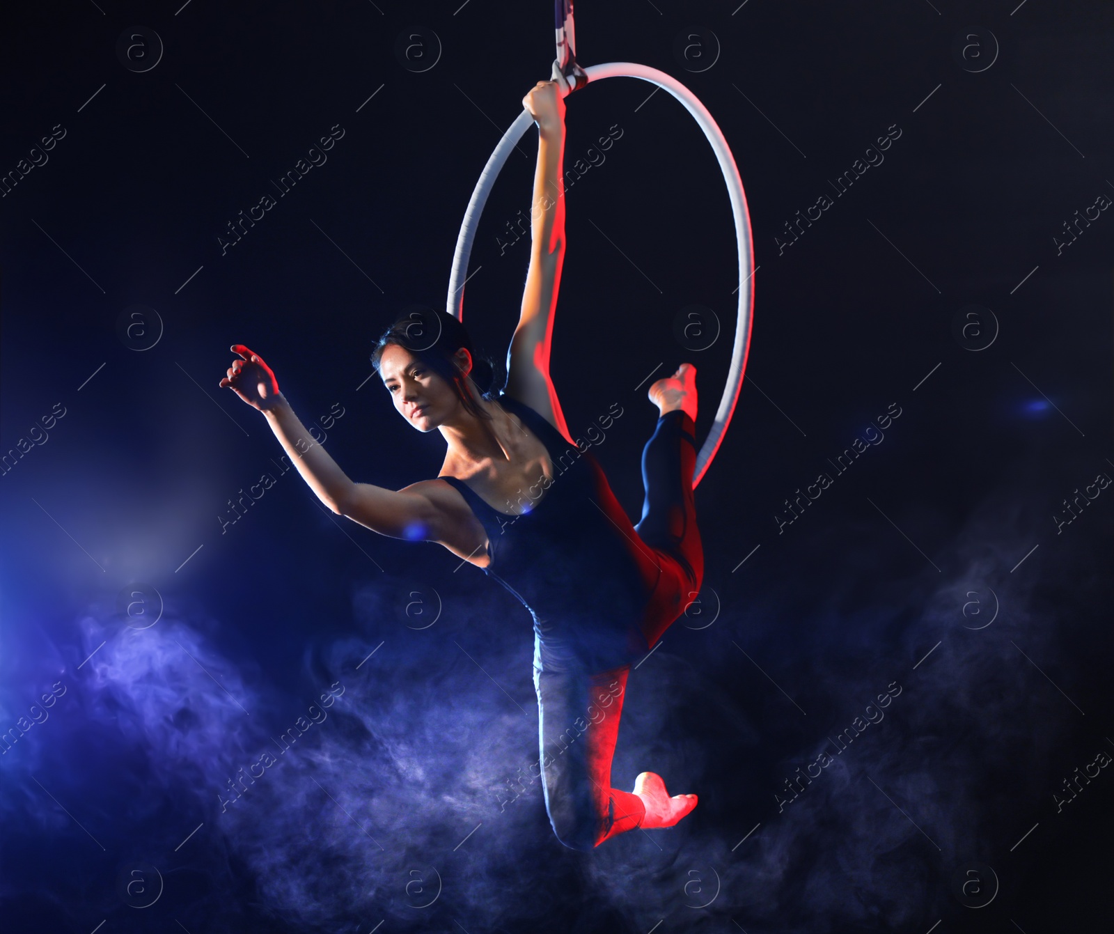 Photo of Young woman performing acrobatic element on aerial ring against dark background