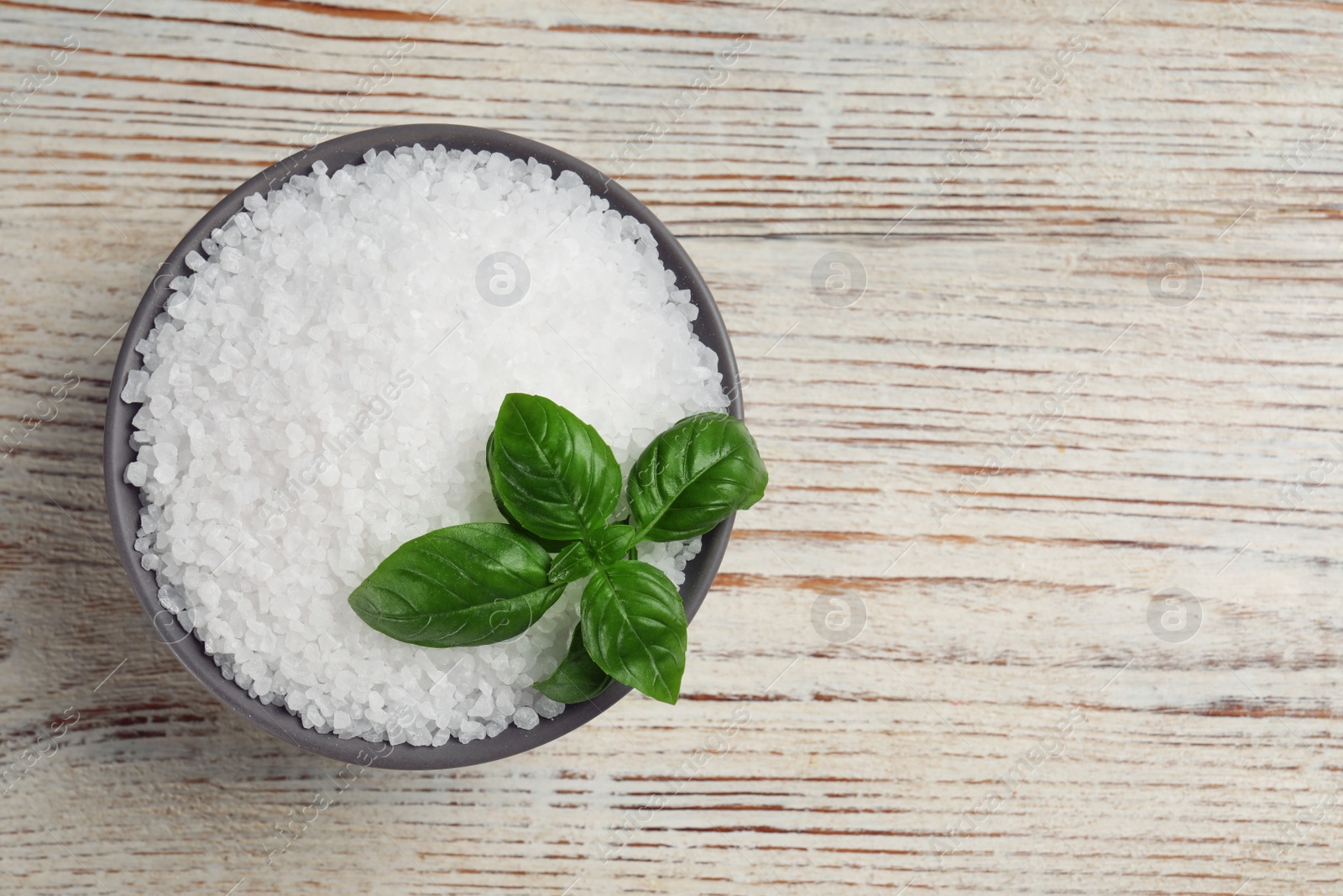 Photo of Bowl of natural sea salt and basil leaf on white wooden table, top view. Space for text