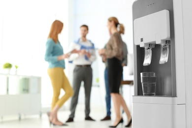 Photo of Modern water cooler with glass and blurred office employees on background. Space for text