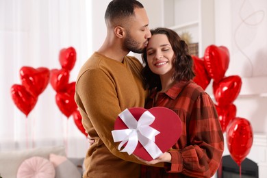 Photo of Happy couple celebrating Valentine's day. Beloved woman with gift box in room decorated with heart shaped air balloons