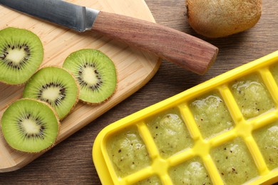 Photo of Kiwi puree in ice cube tray and fresh kiwi fruits on wooden table, flat lay. Ready for freezing