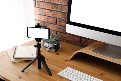 Tripod with smartphone near computer on wooden table indoors. Mockup for design