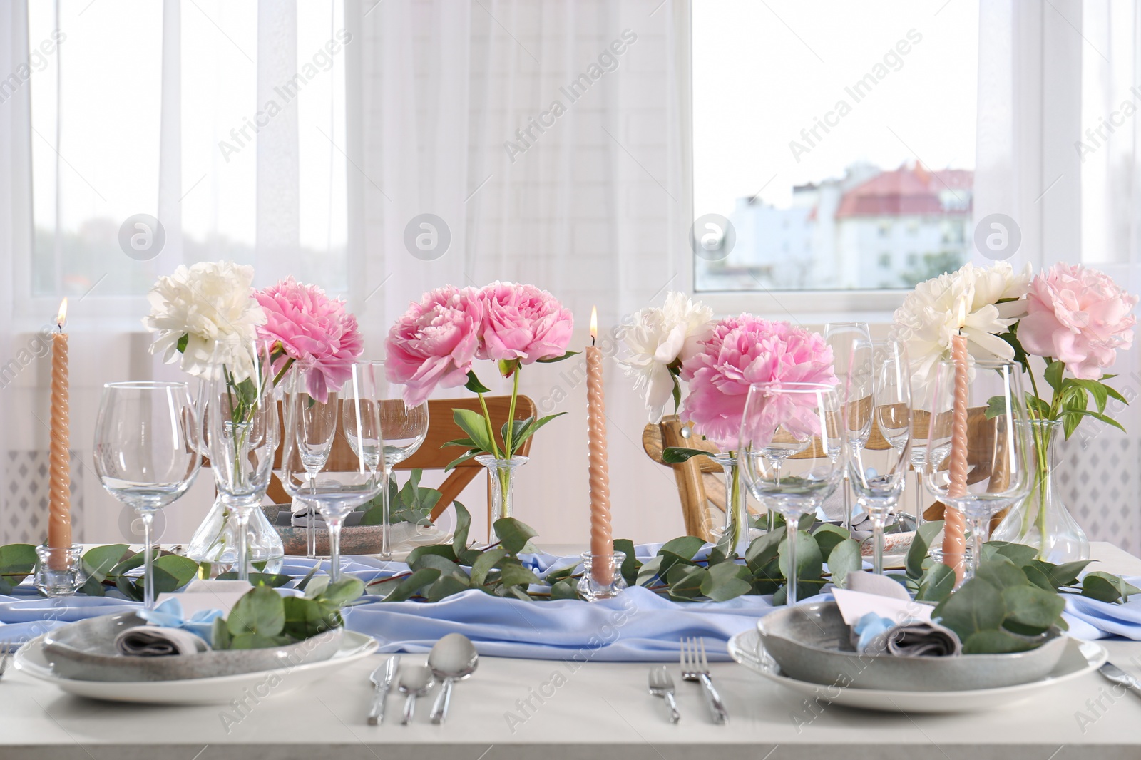 Photo of Beautiful table setting. Plates with greeting cards, napkins and branches near glasses, peonies, burning candles and cutlery on table in room