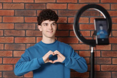 Photo of Smiling teenage blogger making heart gesture to his subscribers while streaming indoors