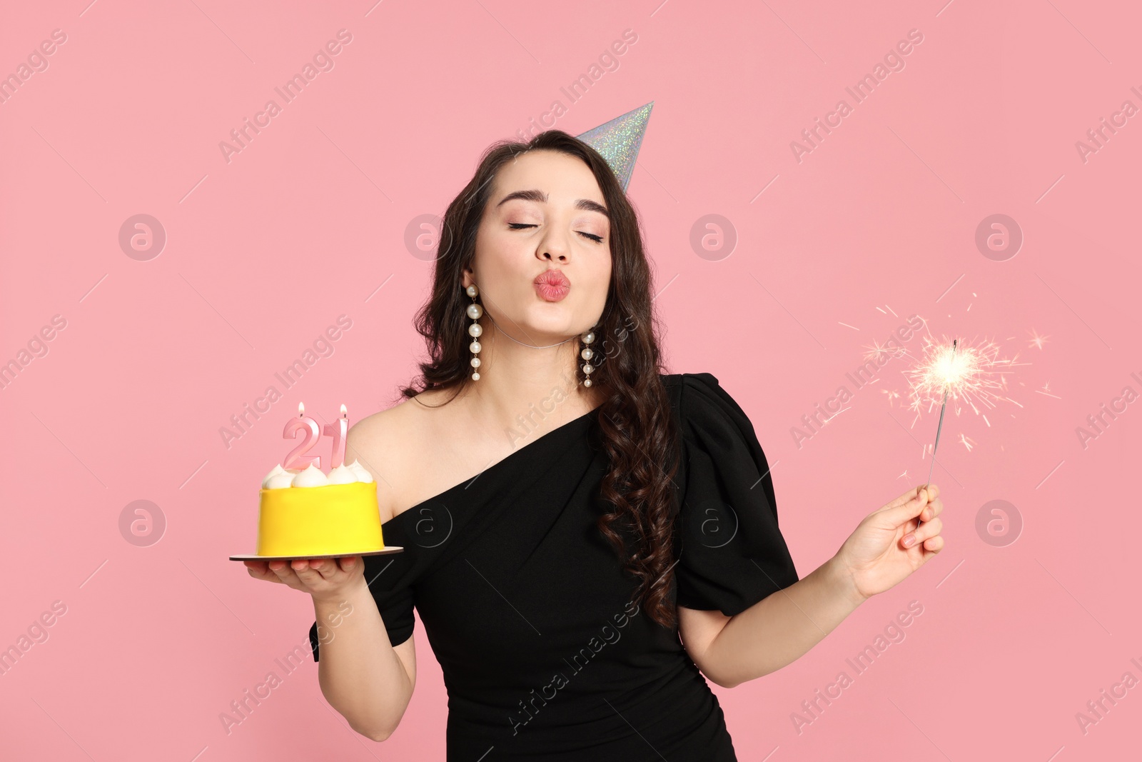 Photo of Coming of age party - 21st birthday. Woman sending air kiss and holding delicious cake with number shaped candles and sparkler on pink background