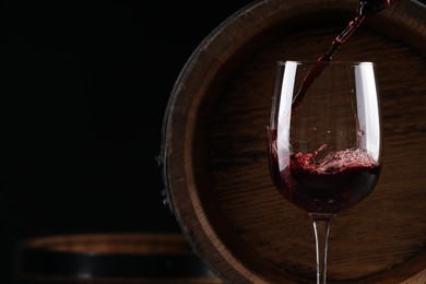 Photo of Pouring red wine into glass near wooden barrel against black background. Space for text