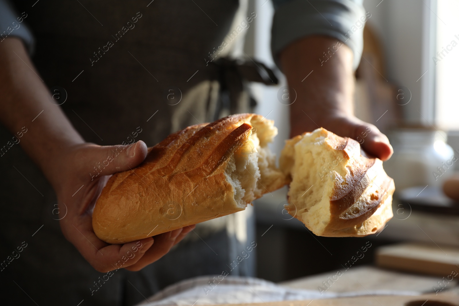 Photo of Man breaking loaf of fresh bread at wooden table indoors, closeup